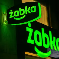 Dwa zielone, podświetlane neony z logo "Żabka" na ciemnej ścianie.