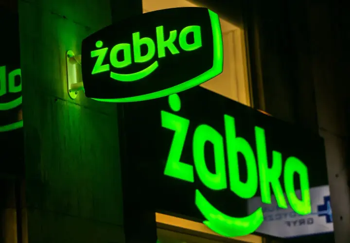 Dwa zielone, podświetlane neony z logo "Żabka" na ciemnej ścianie.