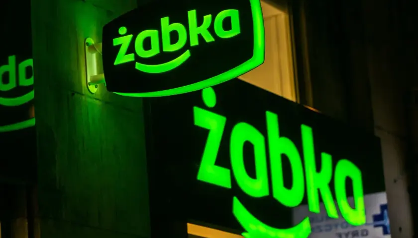 Dwa zielone, podświetlane neony z logo "Żabka" na ciemnej ścianie.